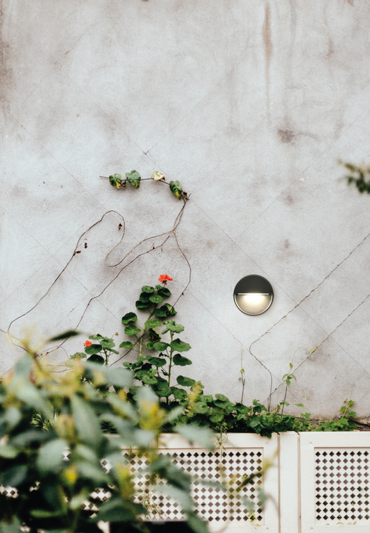 Balise murale extérieure Spica ronde et discrète, diffusant une lumière douce et ciblée idéale pour baliser les zones de passage et structurer les extérieurs avec élégance.